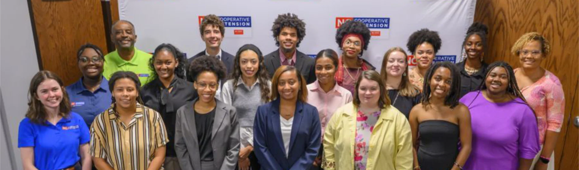 A group of people pose together for a photo in front of a backdrop displaying the NC Cooperative Extension logo. The group includes men and women standing in two rows, smiling at the camera.