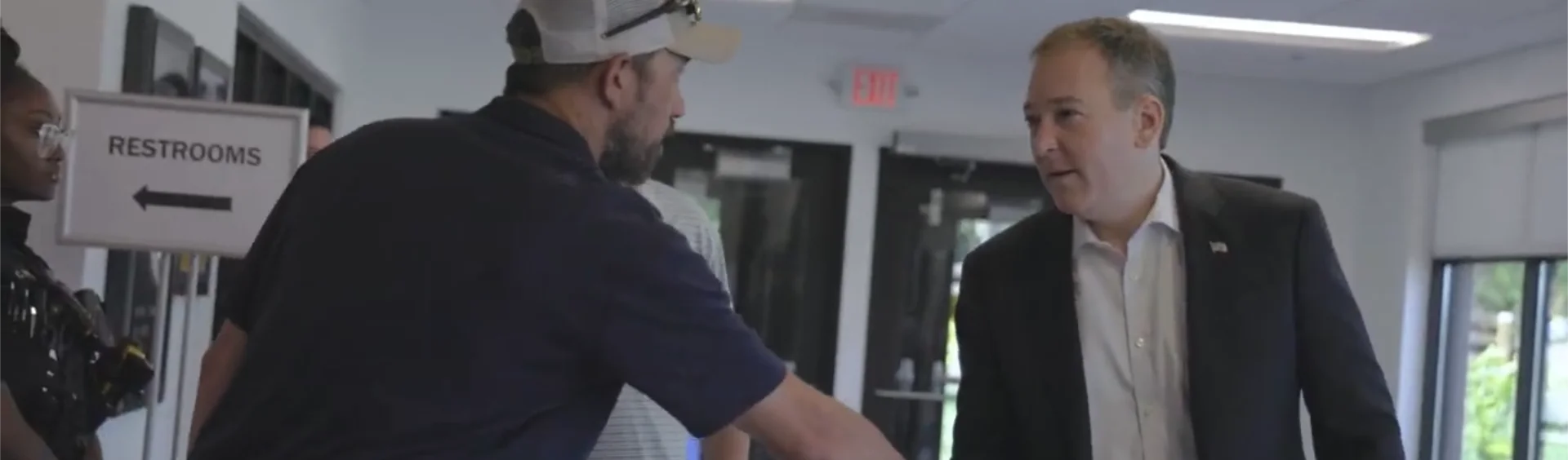 A man in a dark suit shaking hands with a man wearing a dark t-shirt and a baseball cap in an indoor hallway, with a 'Restrooms' sign visible in the background.