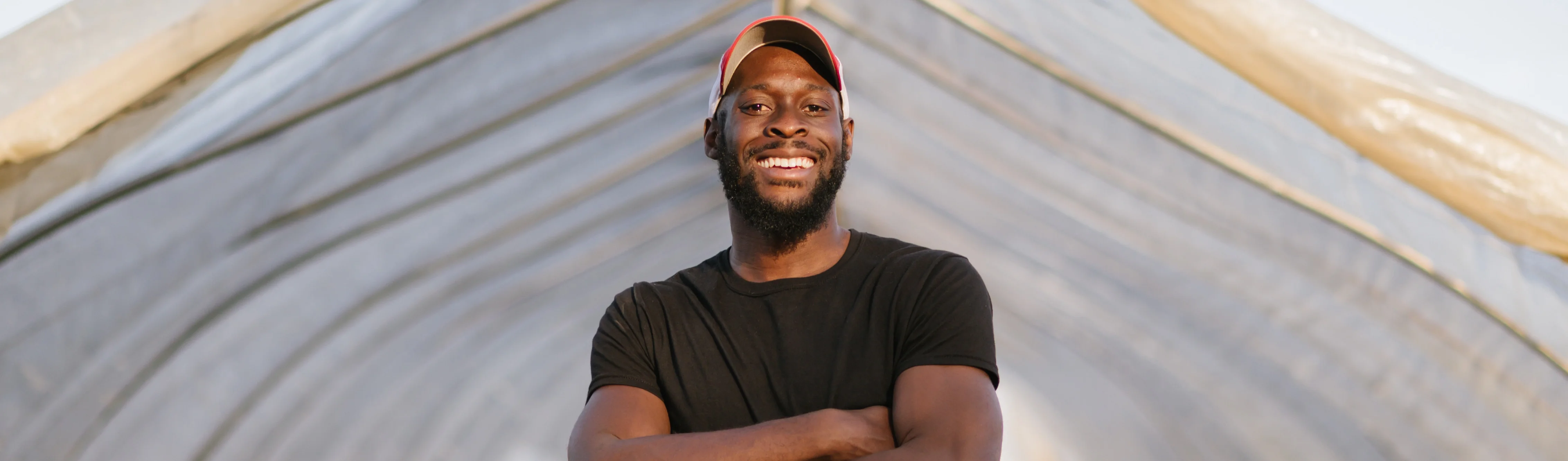 A smiling Black man wearing a black t-shirt and a red and white baseball cap stands with his arms crossed inside a high tunnel greenhouse structure.