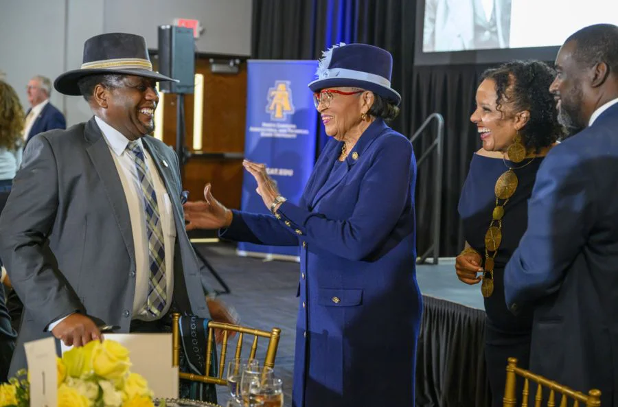 A group of four people stand together at an indoor event, smiling and laughing. A woman in a blue suit and matching hat reaches out warmly toward a man in a gray suit and hat. Two other attendees stand nearby, also smiling. A table with yellow flowers sits in the foreground, and an event banner is visible in the background.